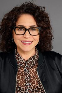 A person with curly brown hair, wearing glasses, a black jacket, and a leopard print shirt, smiles while looking at the camera with the confidence of a trusted advisor.