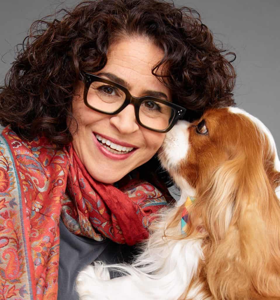 A woman with curly hair and glasses smiles as a Cavalier King Charles Spaniel wearing a colorful scarf touches her face, embodying the joy you'd expect when chatting with an insurance advisor.